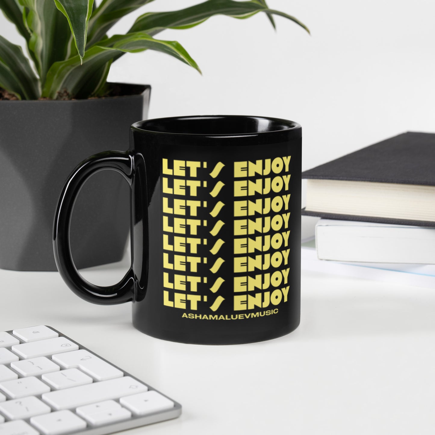 Black mug with yellow text 'LET'S ENJOY' on a desk with a plant and books in the background
