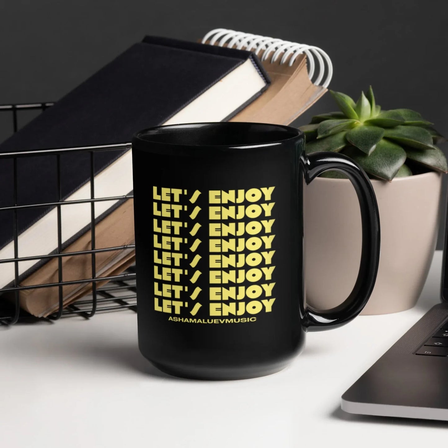 Black mug with yellow text on a desk with a laptop and books
