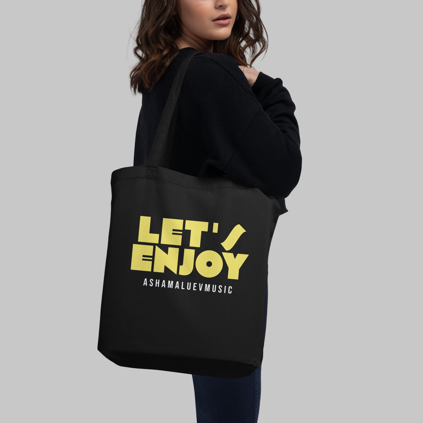 Black tote bag with yellow text 'LET'S ENJOY' and 'ASHAMALUEVMUSIC' and a person holding it on a grey background