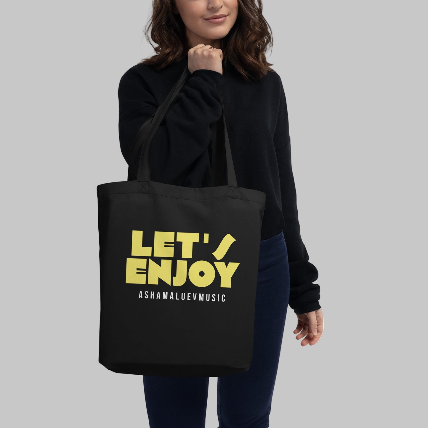 Black tote bag with yellow text 'LET'S ENJOY' and a person holding it on a grey background
