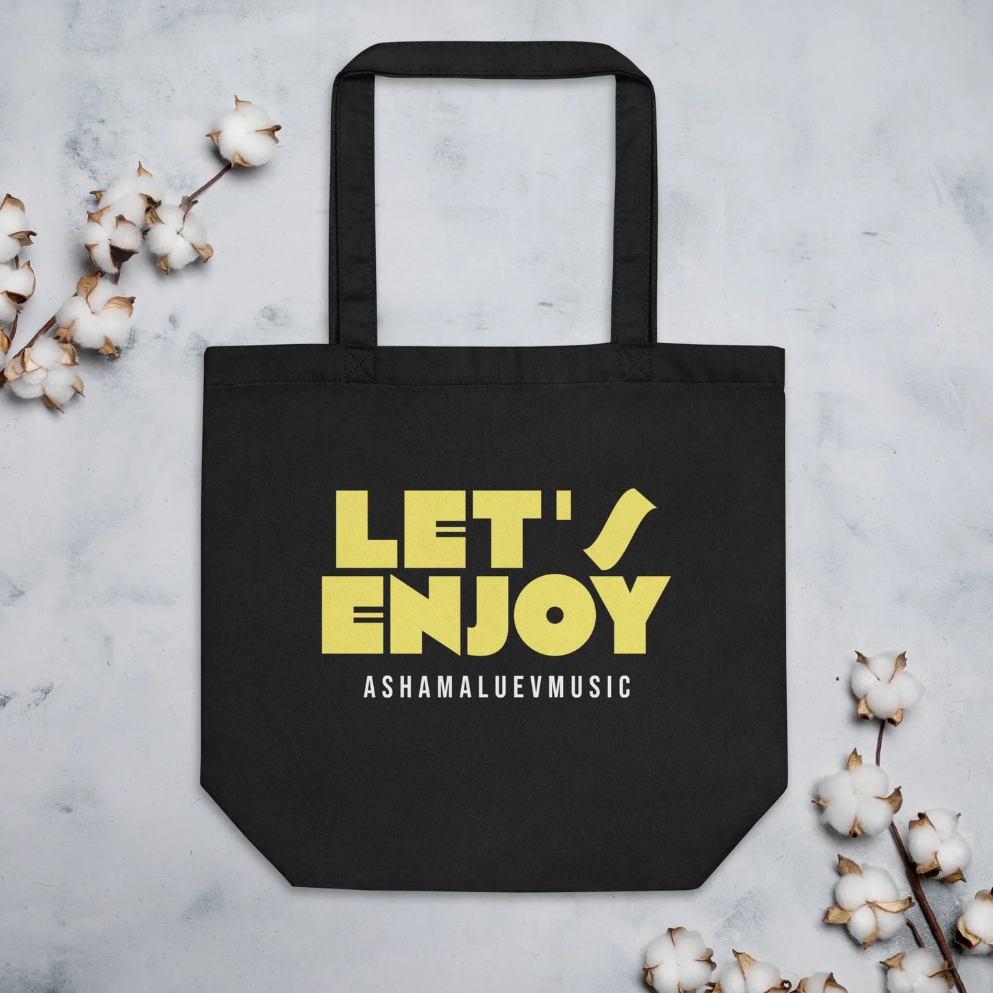 lack tote bag with yellow text 'LET'S ENJOY' and 'ASHAMALUEVMUSIC' on a light gray background with cotton branches.