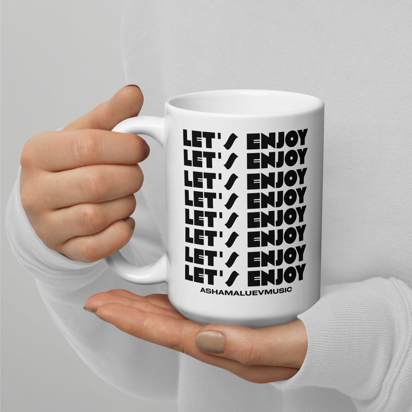 White mug with 'LET'S ENJOY' text held by a person against a gray background