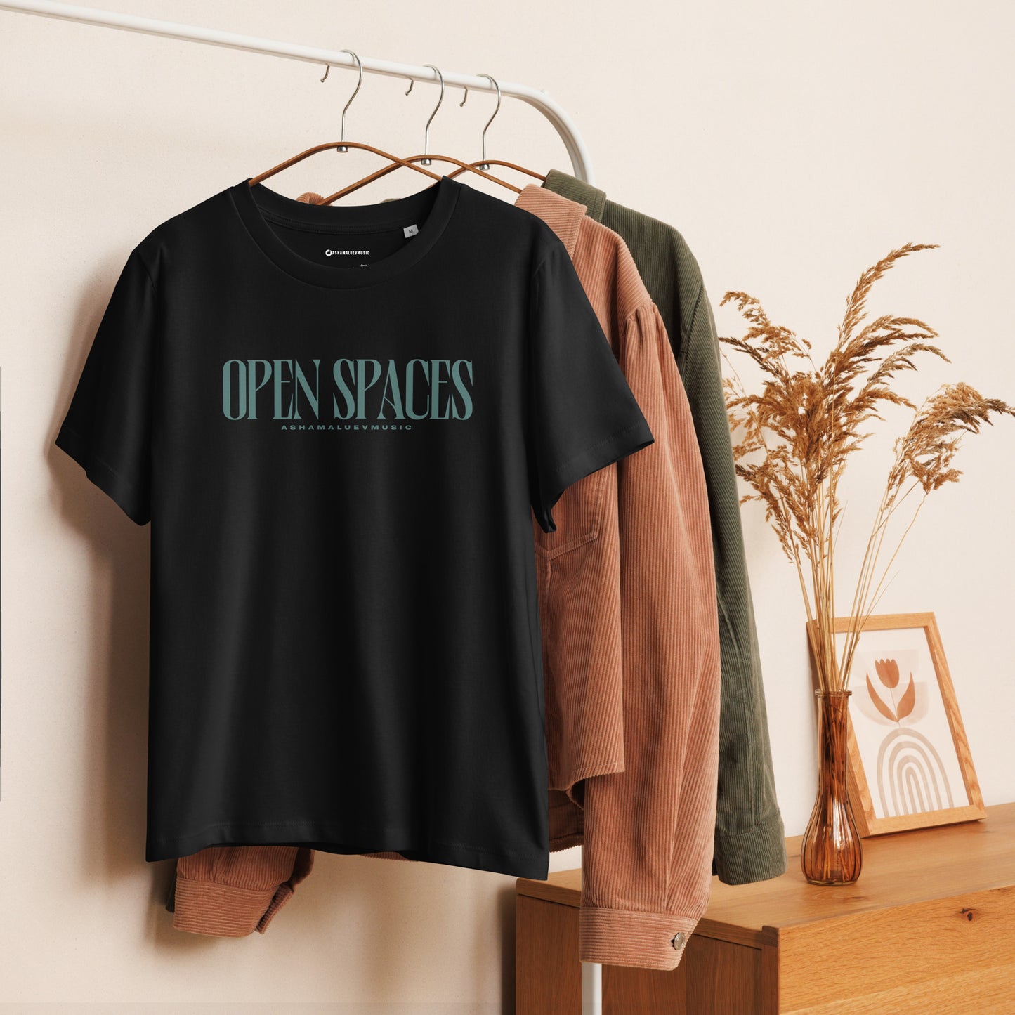 Women's black short sleeve t-shirt with turquoise text 'OPEN SPACES' and 'AShamaluevMusic' hanging on a rack with other clothing items.