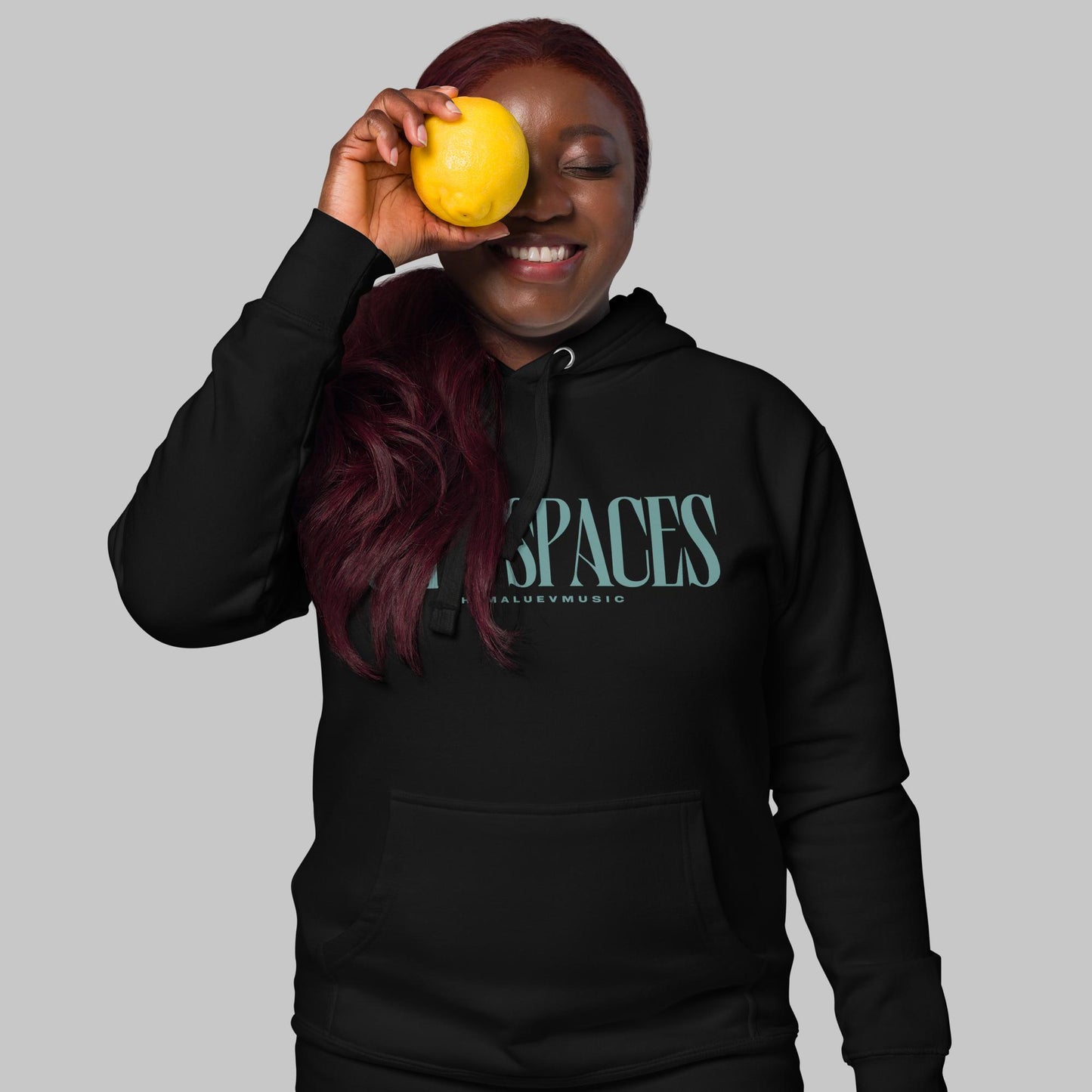 Women wearing a black premium hoodie with turquoise text 'Open Spaces' and 'AShamaluevMusic', holding a yellow fruit against their eye.