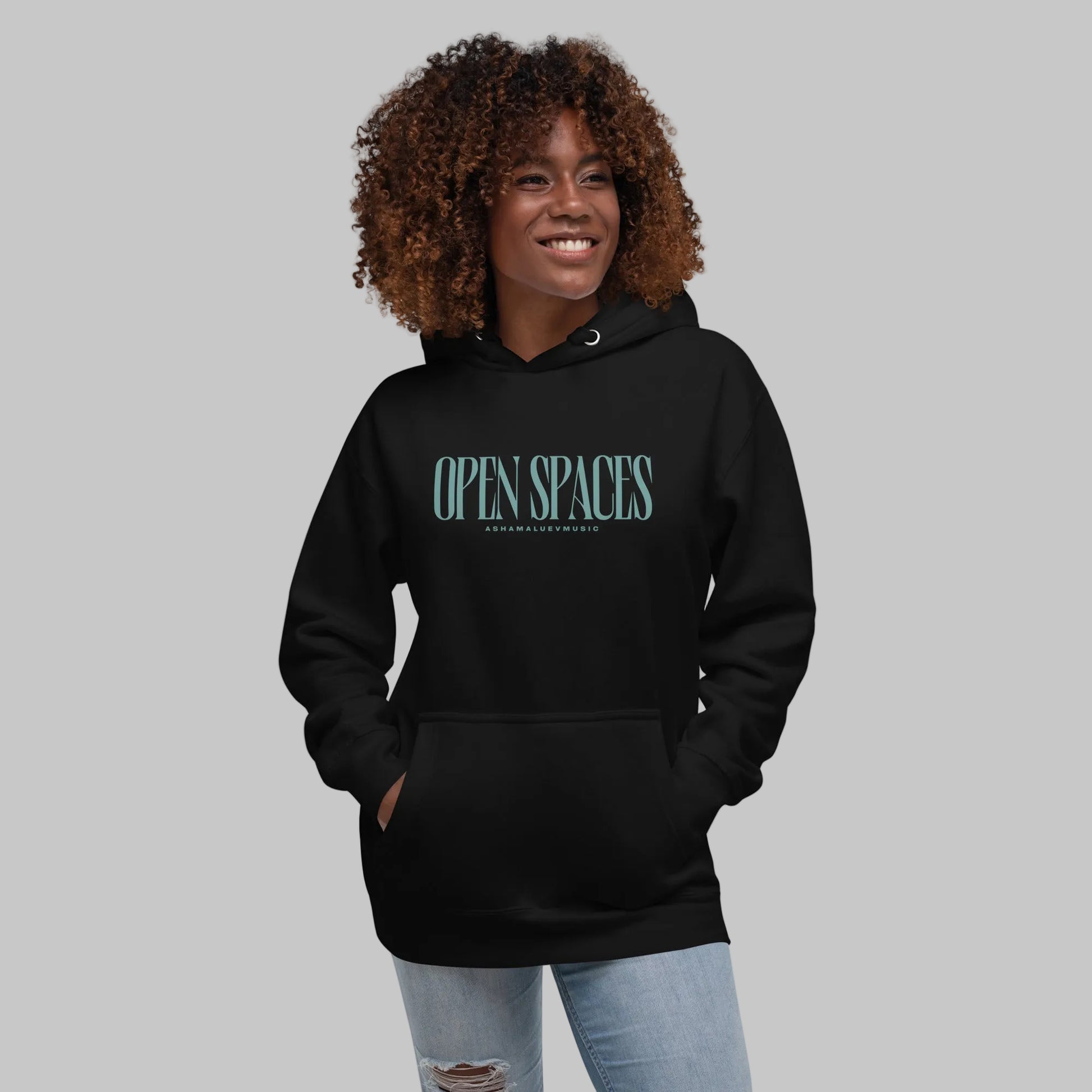 Woman wearing a black premium hoodie with turquoise text 'Open Spaces' and 'AShamaluevMusic' on a gray background.