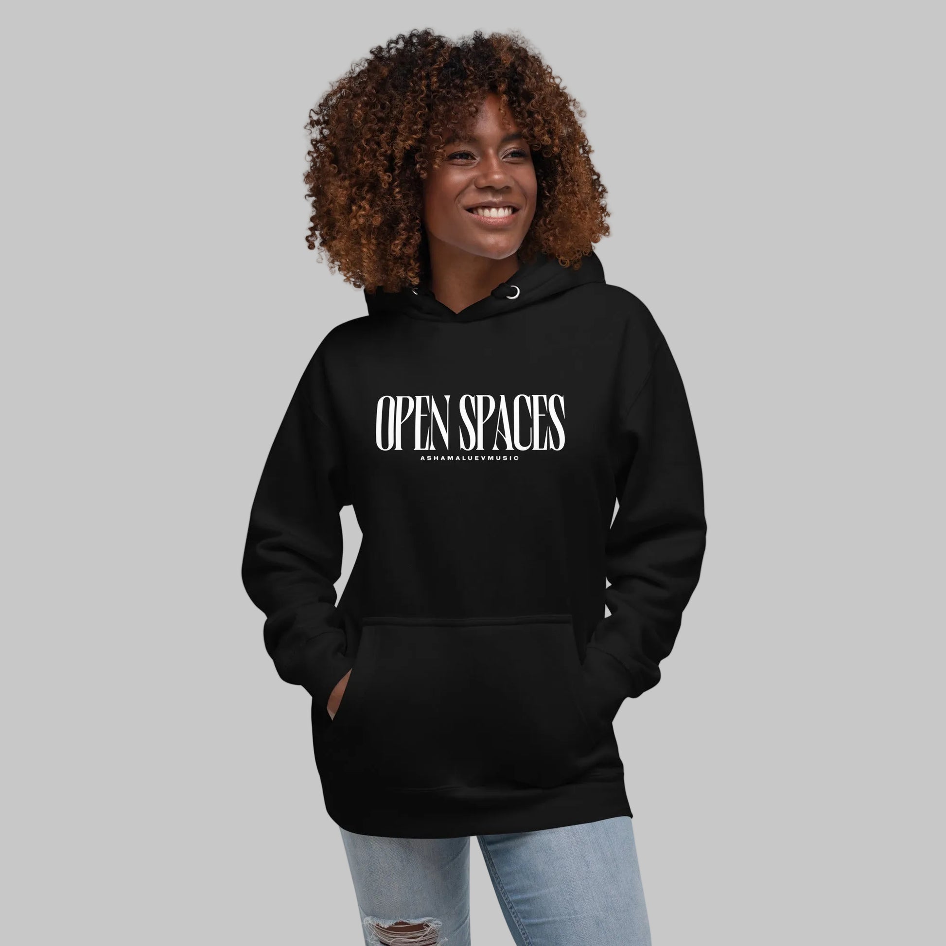 Woman wearing a black unisex pullover hoodie with white text 'Open Spaces' and 'AShamaluevMusic' on a gray background.