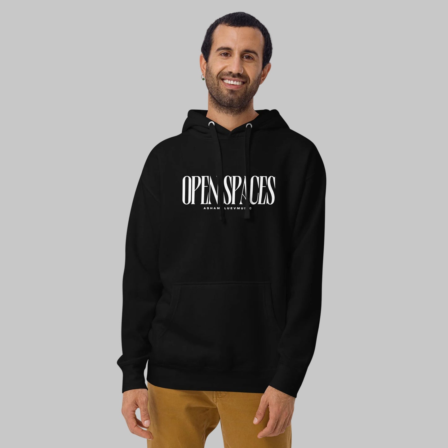 Man wearing a black unisex pullover hoodie with white text 'Open Spaces' and 'AShamaluevMusic' on a gray background.