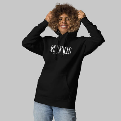 Woman wearing a black unisex pullover hoodie with white text 'Open Spaces' and 'AShamaluevMusic' on a gray background.