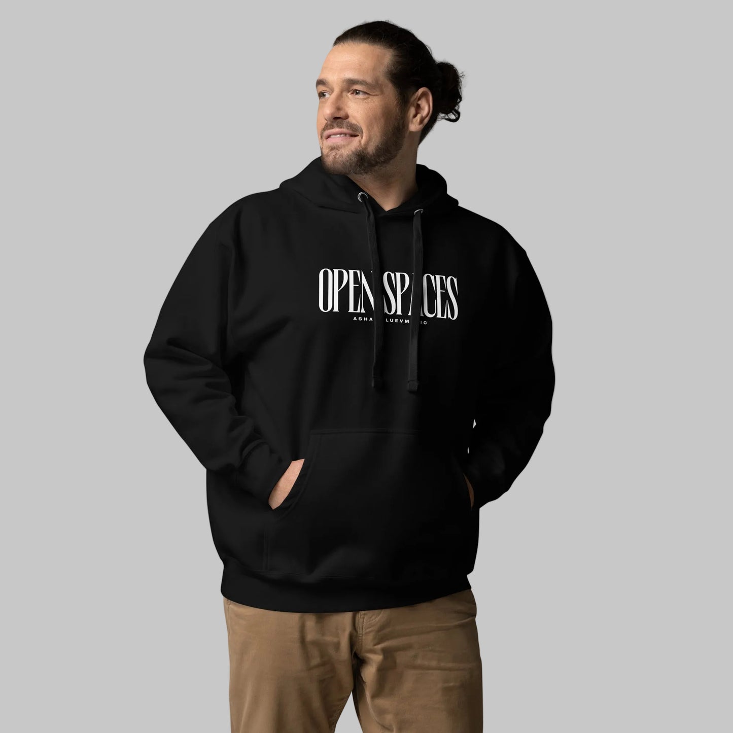 Man wearing a black unisex pullover hoodie with white text 'Open Spaces' and 'AShamaluevMusic' on a gray background.