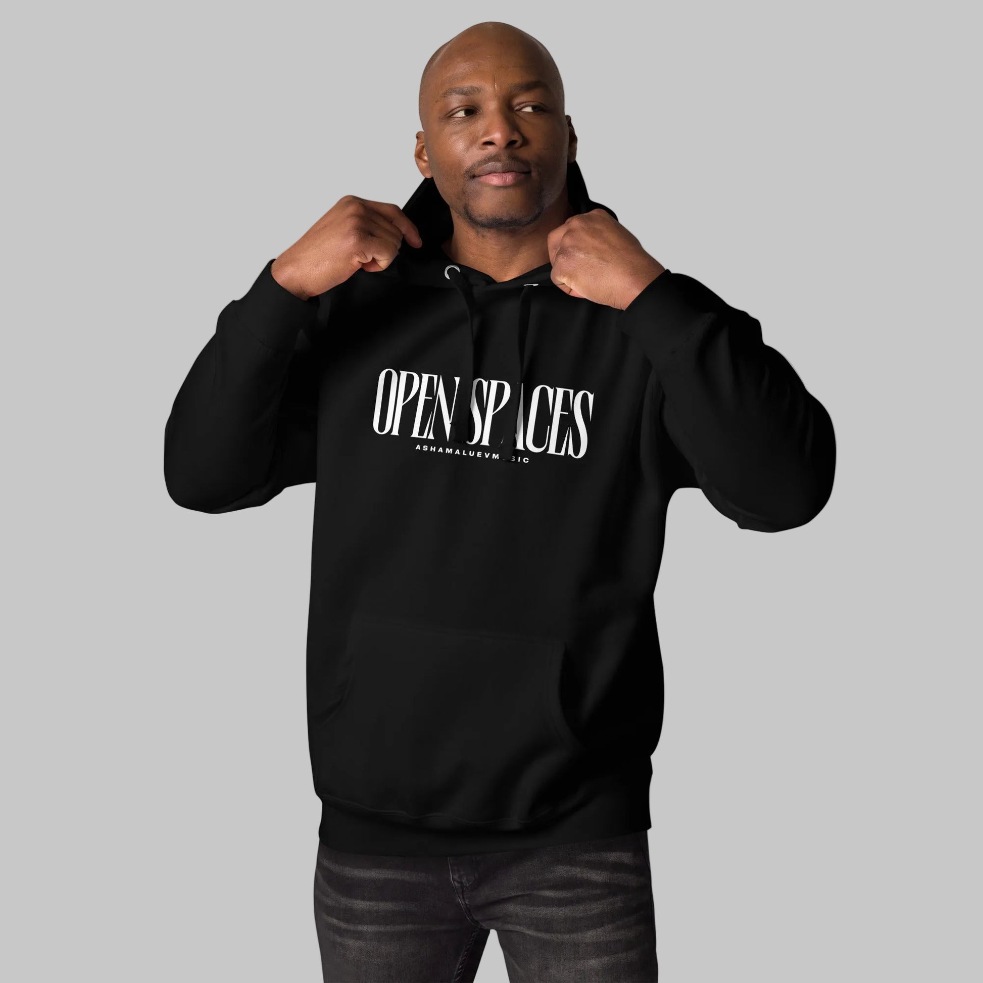 Man wearing a black unisex pullover hoodie with white text 'Open Spaces' and 'AShamaluevMusic' on a gray background.