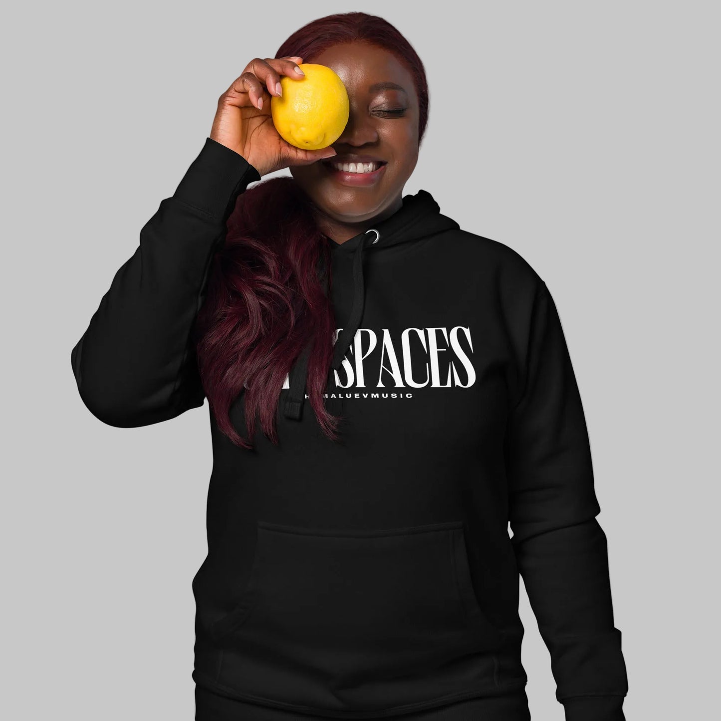 Woman wearing a black unisex pullover hoodie with white text 'OPEN SPACES' and 'AShamaluevMusic', holding a yellow fruit against a gray background