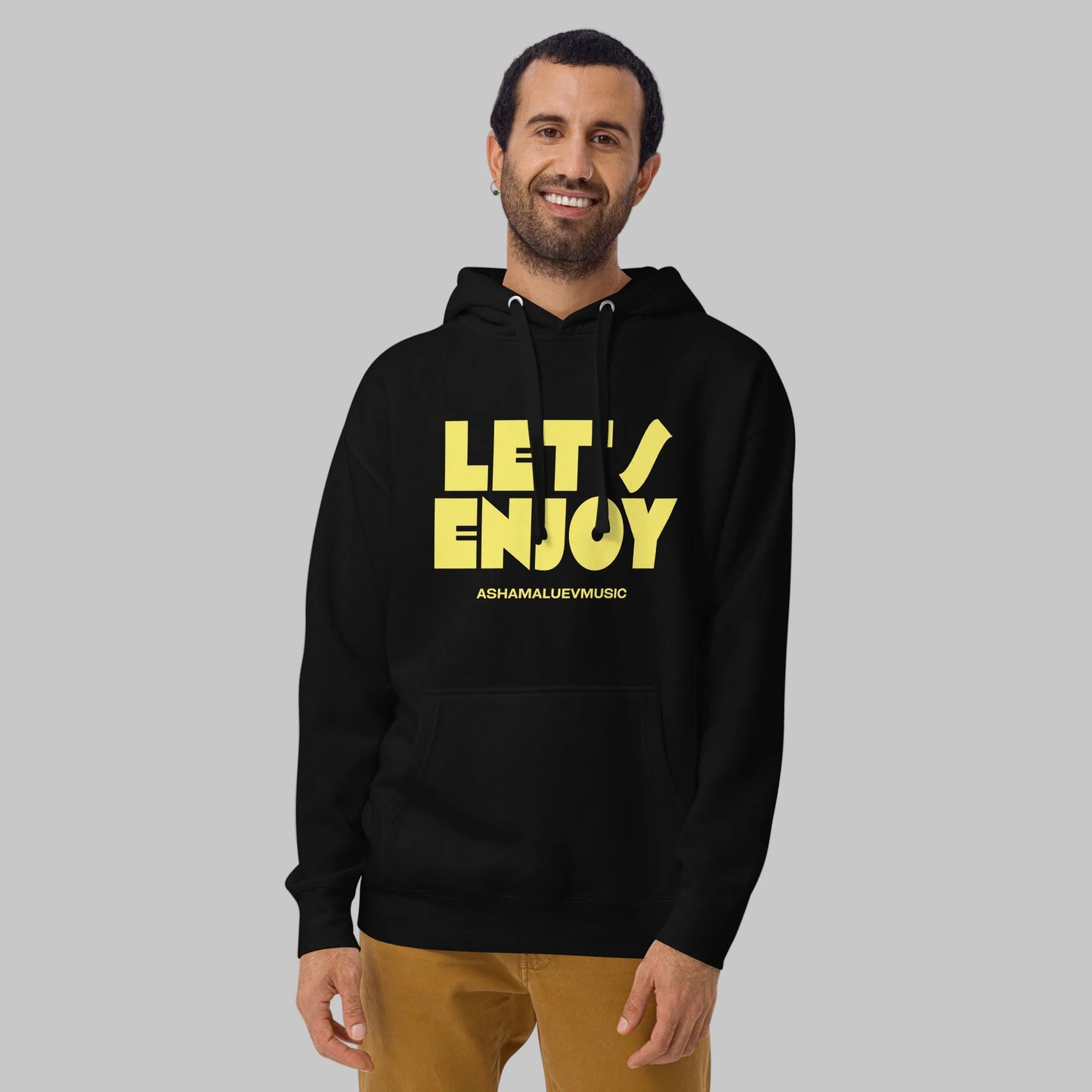 Man wearing a black unisex pullover hoodie with yellow text 'Let's Enjoy' and 'AShamaluevMusic' on a gray background.