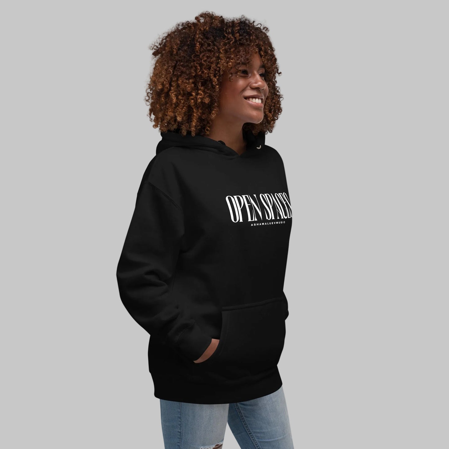 Woman wearing a black unisex pullover hoodie with white text 'Open Spaces' and 'AShamaluevMusic' on a gray background.