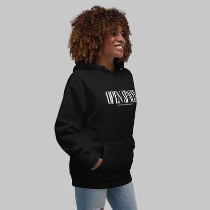 Woman wearing a black unisex pullover hoodie with white text 'Open Spaces' and 'AShamaluevMusic' on a gray background.