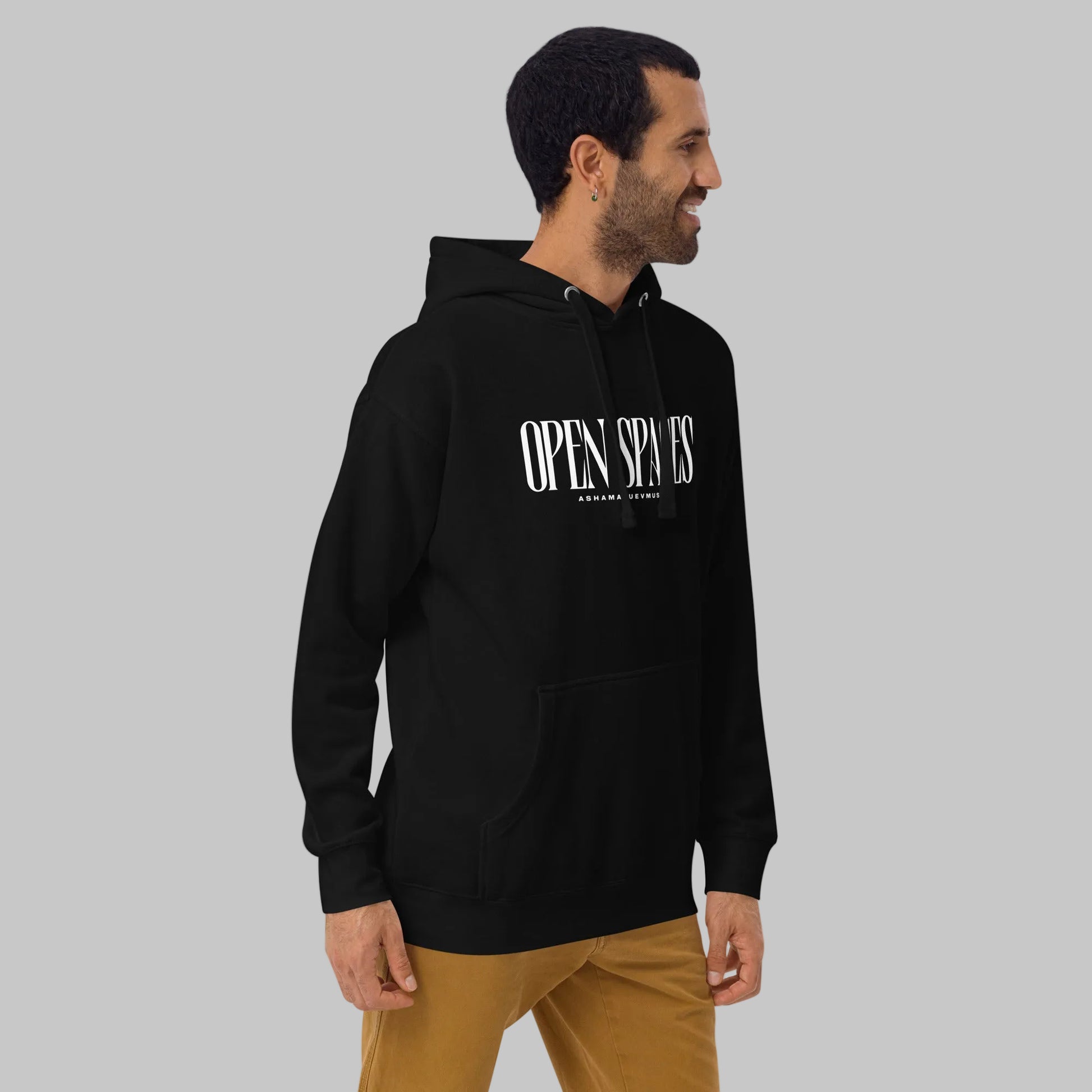 Man wearing a black unisex pullover hoodie with white text 'Open Spaces' and 'AShamaluevMusic' on a gray background.