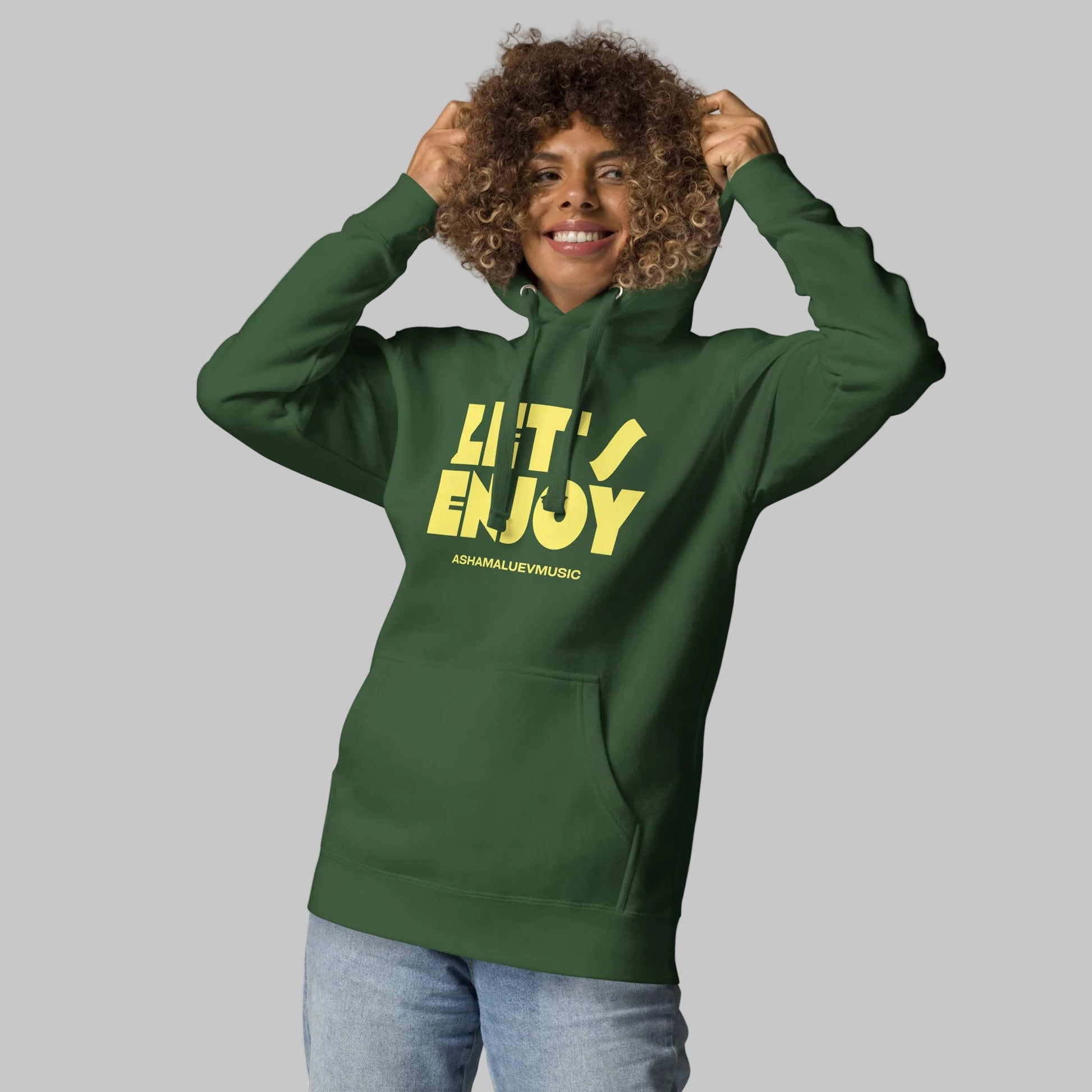 Woman wearing a green unisex pullover hoodie with yellow text 'Let's Enjoy' and 'AShamaluevMusic' on a gray background.