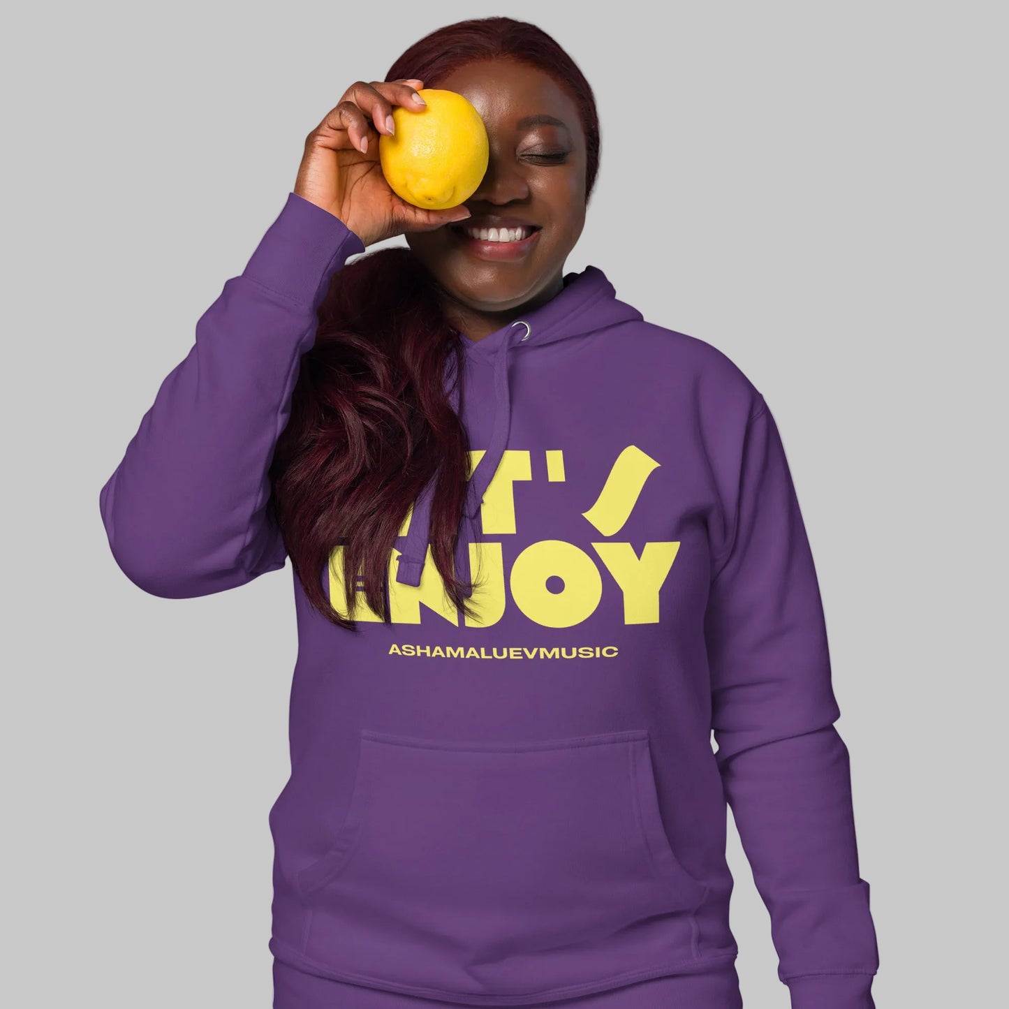 Woman wearing a purple unisex pullover hoodie with yellow text 'Let's Enjoy' and 'AShamaluevMusic' on a gray background.