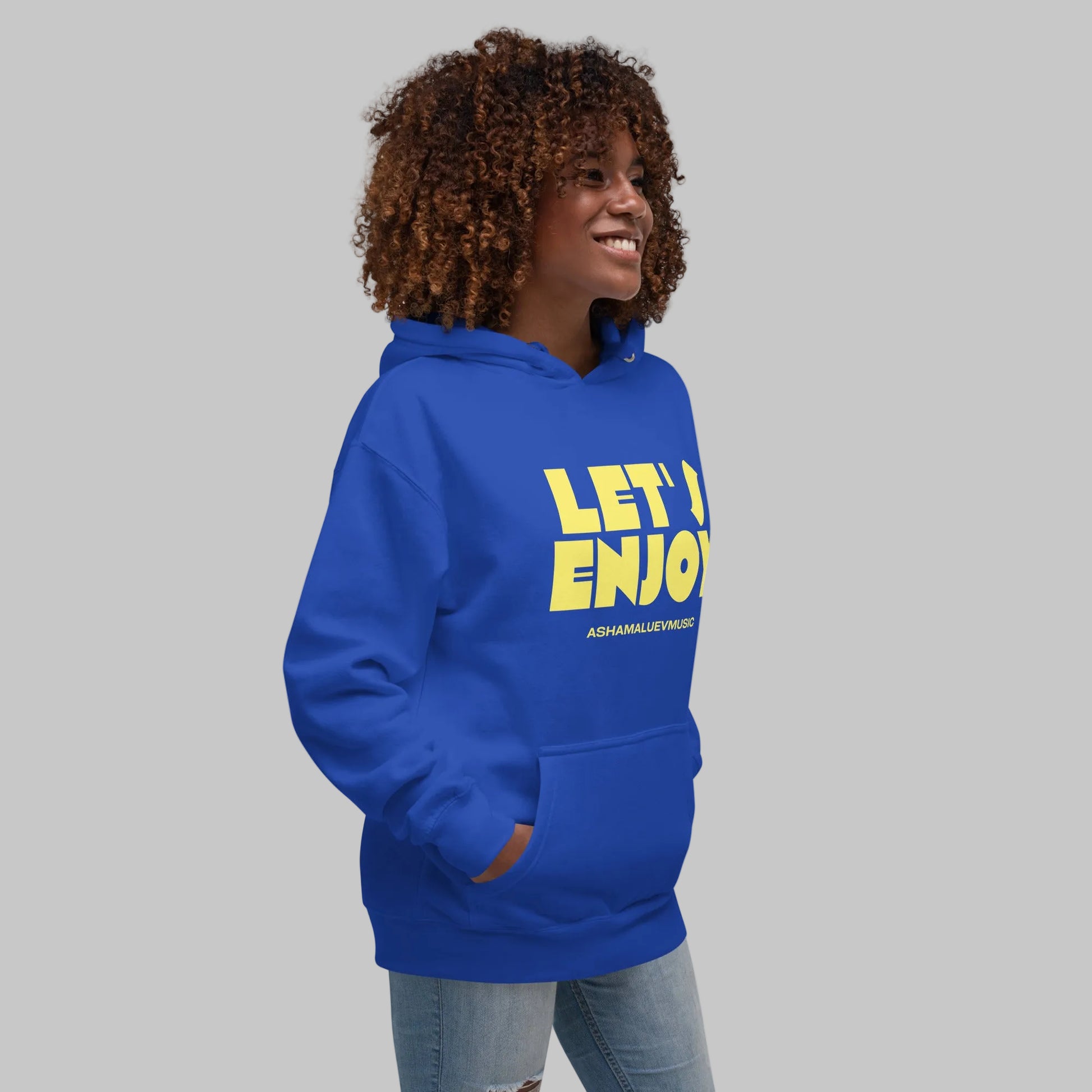 Woman wearing a blue unisex pullover hoodie with yellow text 'Let's Enjoy' and 'AShamaluevMusic' on a gray background.