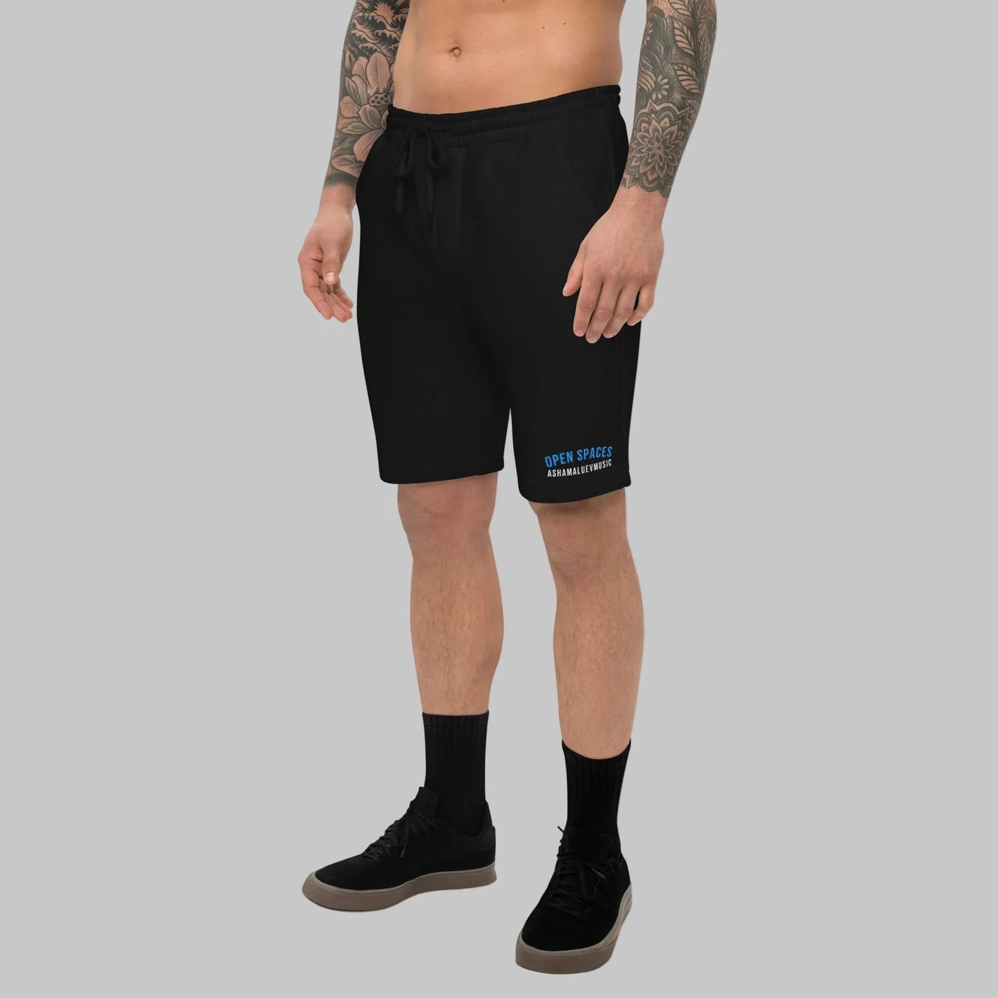 Man wearing black fleece shorts with 'Open Spaces' and 'AShamaluevMusic' embroidered on a gray background