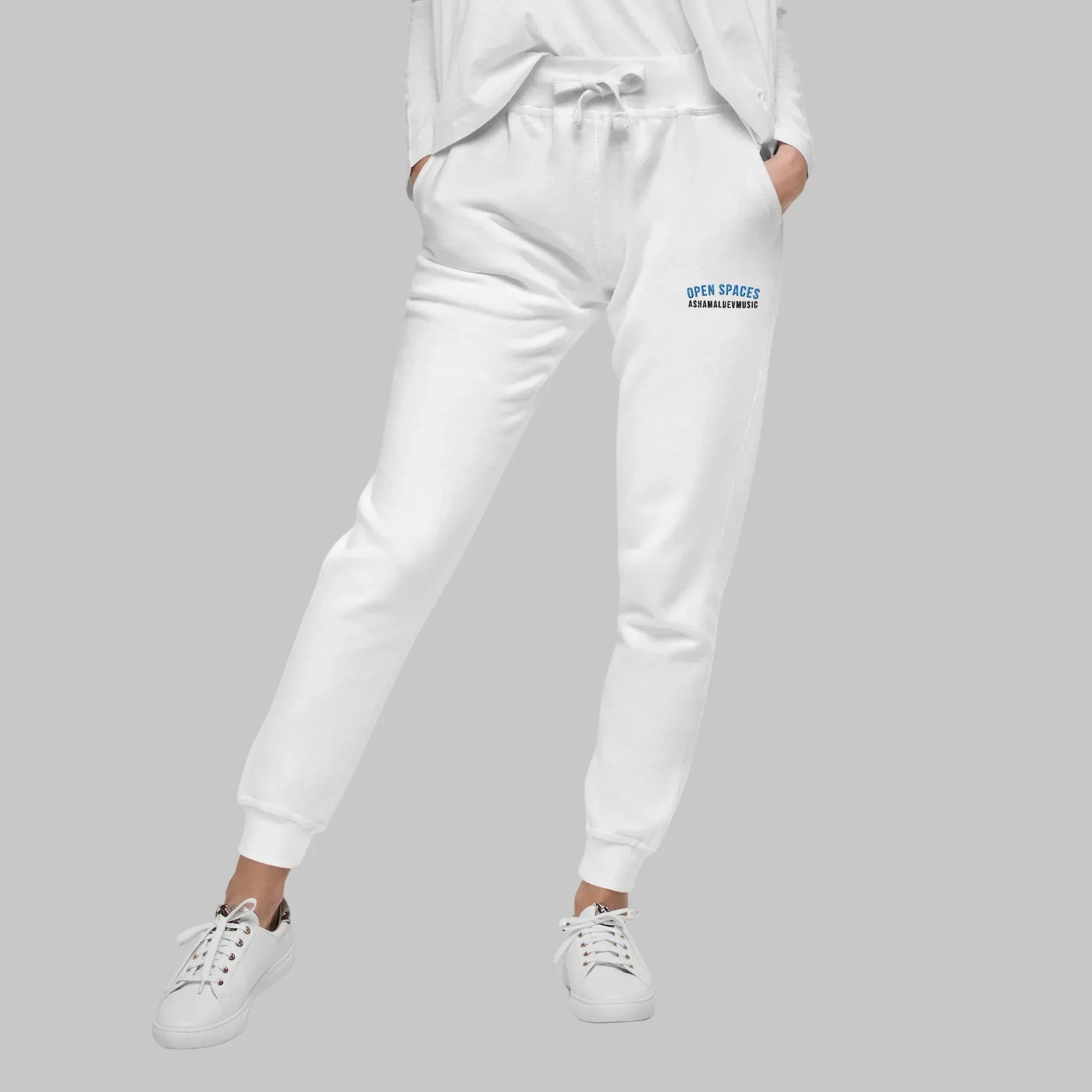 Woman wearing white sweatpants with 'Open Spaces' and 'AShamaluevMusic' embroidered on a gray background